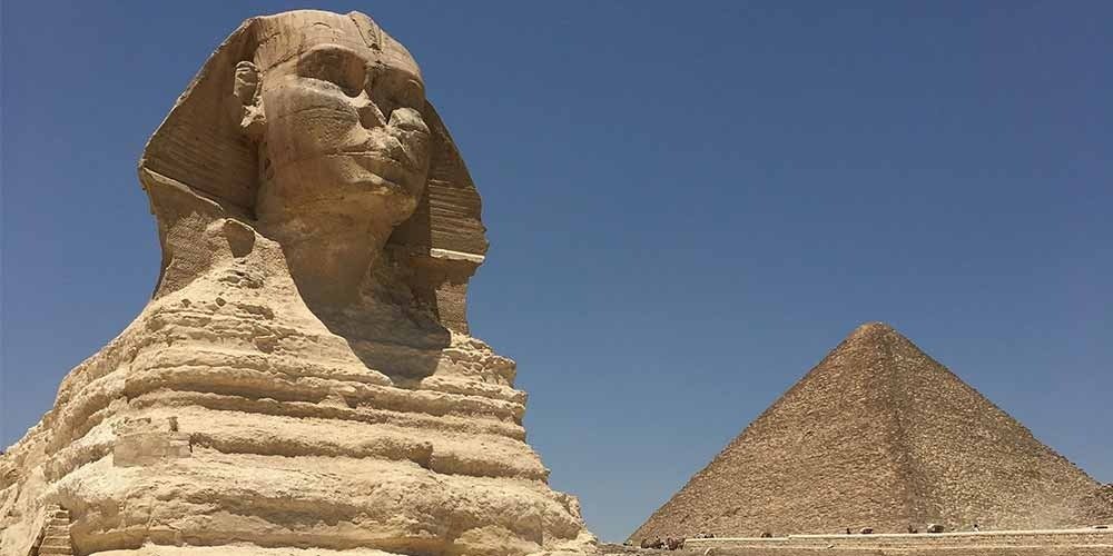 A majestic low-angle side profile of the Sphinx carved from weathered limestone, showing its facial features and royal nemes headcloth. The massive monument stands in the foreground, while the apex of a Great Pyramid rises behind it under a vast, clear blue sky