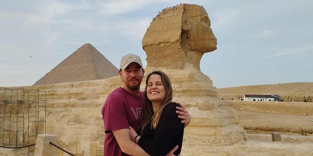 A happy man and woman embracing and smiling in front of the side profile of the Sphinx under a soft evening sky. The colossal monument stands on its stone base with birds perched on its head, while a Great Pyramid and a tourist bus are visible in the background.