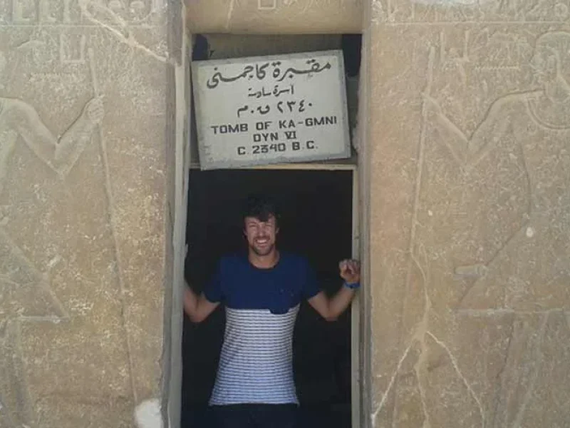 A tourist taking a photo inside the tomb of Kagmni at Saqqara, showing ancient Egyptian wall reliefs.