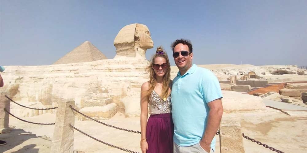 Un couple souriant pose devant le Grand Sphinx de Gizeh avec la Grande Pyramide en arrière-plan lors d'une excursion Pyramides depuis Sharm El-Sheikh