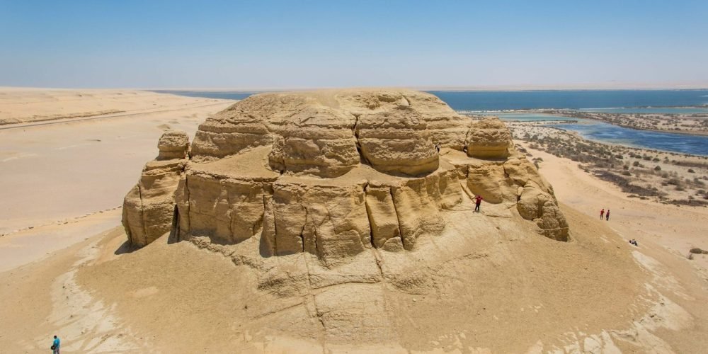 A high-angle landscape shot of Mudawara Mountain in the Fayoum desert. The mountain is a massive, rounded rock formation made of light brown sandstone with visible horizontal layers and vertical cracks. Several small figures of tourists are visible climbing the slopes and standing on the ridges. In the background, the deep blue waters of Magic Lake stretch toward the horizon, bordered by sandy shores and green patches of vegetation. The scene is captured under a bright, clear blue sky during a Fayoum sharing tour