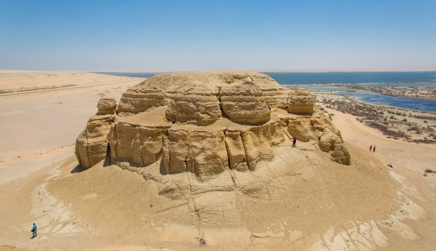 A high-angle landscape shot of Mudawara Mountain in the Fayoum desert. The mountain is a massive, rounded rock formation made of light brown sandstone with visible horizontal layers and vertical cracks. Several small figures of tourists are visible climbing the slopes and standing on the ridges. In the background, the deep blue waters of Magic Lake stretch toward the horizon, bordered by sandy shores and green patches of vegetation. The scene is captured under a bright, clear blue sky during a Fayoum sharing tour