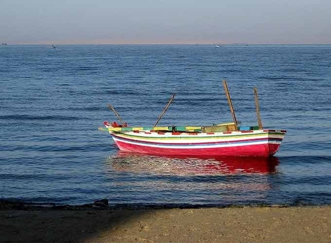 A horizontal eye-level shot of a small, traditional wooden rowboat floating on the calm, deep blue water of Lake Qarun. The boat is painted with vibrant horizontal stripes of pink, white, green, and blue. Two thin wooden masts or oars stick up from the center of the boat. In the far distance, a thin strip of desert coastline is visible under a soft, hazy sky. The foreground shows a small patch of sandy shoreline in the shade. This peaceful scene is captured during a Fayoum sharing tour
