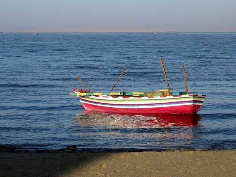 A horizontal eye-level shot of a small, traditional wooden rowboat floating on the calm, deep blue water of Lake Qarun. The boat is painted with vibrant horizontal stripes of pink, white, green, and blue. Two thin wooden masts or oars stick up from the center of the boat. In the far distance, a thin strip of desert coastline is visible under a soft, hazy sky. The foreground shows a small patch of sandy shoreline in the shade. This peaceful scene is captured during a Fayoum sharing tour