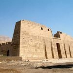 Une vue imposante du grand pylône en pierre d'un temple antique avec des gravures massives de pharaons et de divinités sur les murs. La structure présente des ouvertures verticales étroites et une grande porte centrale, le tout sous un ciel bleu dégagé lors d'un Forfait au Caire et à Louxor Assouan
