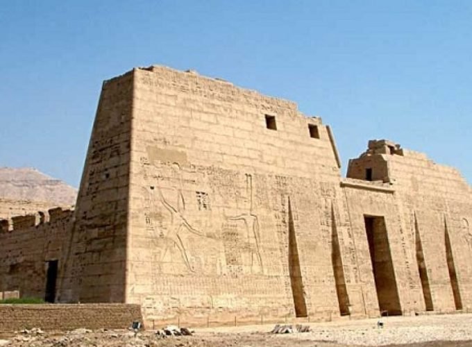 Une vue imposante du grand pylône en pierre d'un temple antique avec des gravures massives de pharaons et de divinités sur les murs. La structure présente des ouvertures verticales étroites et une grande porte centrale, le tout sous un ciel bleu dégagé lors d'un Forfait au Caire et à Louxor Assouan