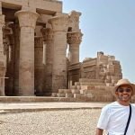 Un jeune homme et une jeune femme souriants، portant des chapeaux de paille et des lunettes de soleil، posent devant les grandes colonnes de pierre du temple de Kom Ombo. Le temple antique en arrière-plan présente des chapiteaux sculptés sous un ciel clair lors d'un Forfait au Caire et à Louxor Assouan