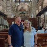 Un homme en t-shirt bleu et une femme blonde en t-shirt blanc posent ensemble à l'intérieur d'une église ancienne. Derrière eux, on voit des rangées de bancs en bois, des piliers en pierre, des murs en briques rouges et une structure d'autel richement décorée sous des lustres allumés lors d'un Forfait au Caire et à Louxor Assouan