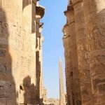 Une rangée de colonnes de pierre géantes et massives couvertes de hiéroglyphes et de bas-reliefs antiques sculptés. Les colonnes se dressent vers un ciel bleu clair, et au loin, on aperçoit la pointe d'un obélisque égyptien s'élevant entre les structures lors d'un Forfait au Caire et à Louxor Assouan