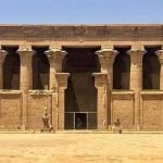 Une vue de la cour intérieure d'un temple antique en pierre avec une colonnade imposante. Les chapiteaux des colonnes sont sculptés en forme de fleurs de papyrus. Au centre, une entrée sombre mène vers l'intérieur du sanctuaire, avec des statues d'Horus en forme de faucon devant la façade lors d'un Forfait au Caire et à Louxor Assouan