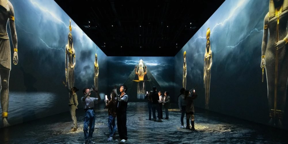 A group of tourists taking photos inside a dark immersive digital room featuring massive wall-to-wall projections of ancient Egyptian gods. The glowing figures stand tall against a background of lightning and stormy skies during a Giza Pyramids & Grand Egyptian Museum & tour