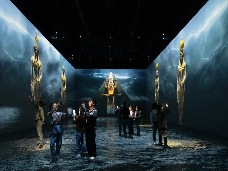 A group of tourists taking photos inside a dark immersive digital room featuring massive wall-to-wall projections of ancient Egyptian gods. The glowing figures stand tall against a background of lightning and stormy skies during a Giza Pyramids & Grand Egyptian Museum & tour