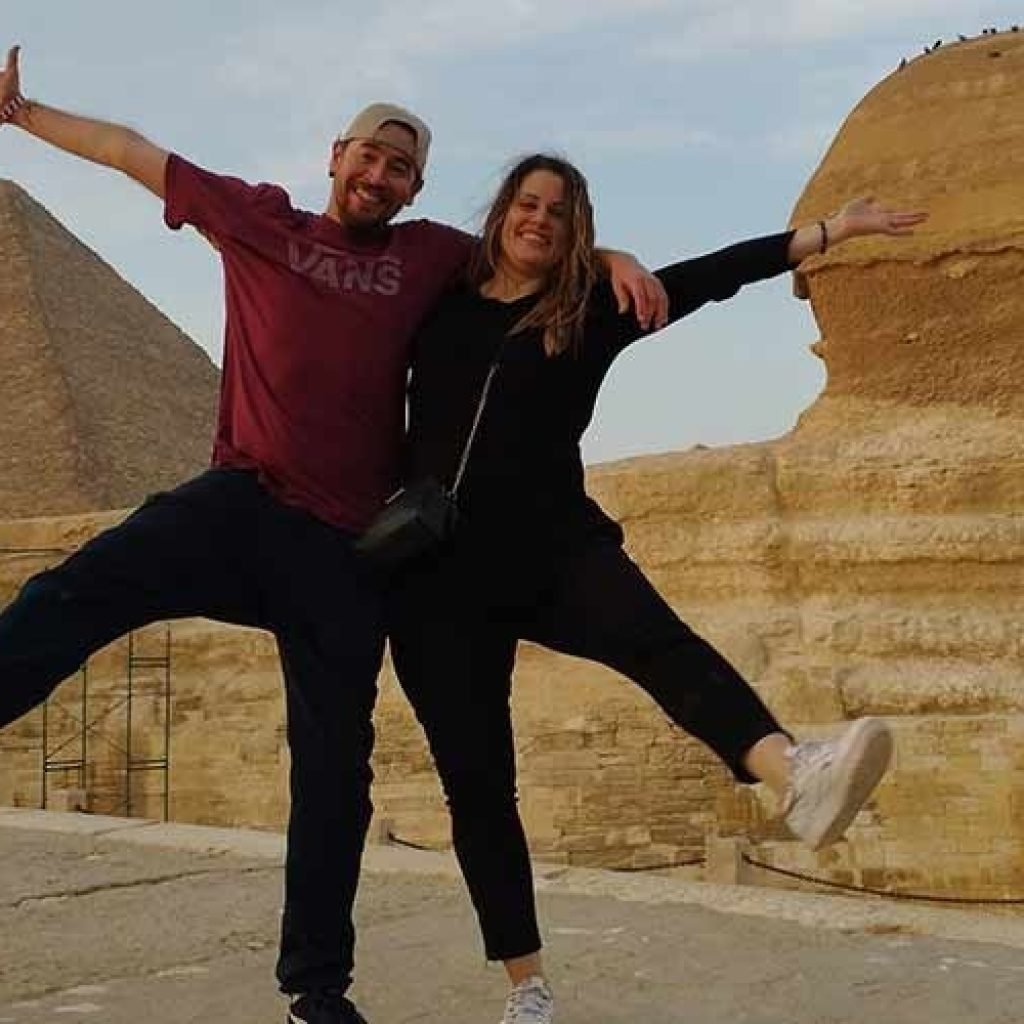 A man in a red Vans t-shirt and a woman in black clothing pose joyfully with arms outstretched in front of the Great Sphinx and a large pyramid at Giza.