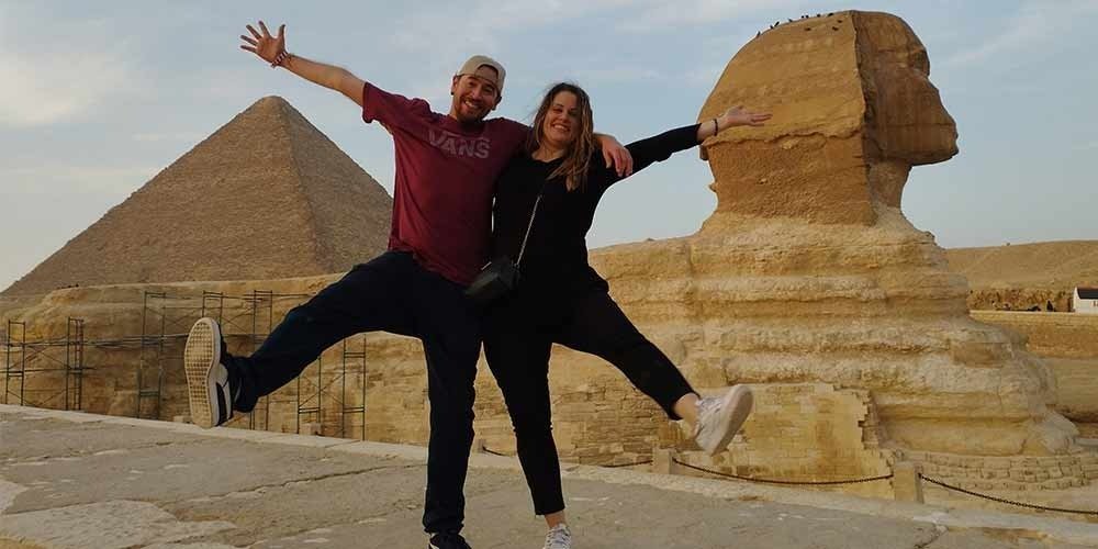 A man in a red Vans t-shirt and a woman in black clothing pose joyfully with arms outstretched in front of the Great Sphinx and a large pyramid at Giza