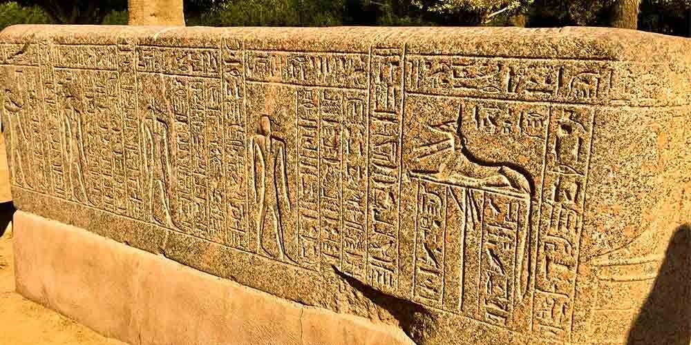 A close-up shot of a large ancient stone monument featuring deeply carved hieroglyphics and figures, including the jackal-headed god Anubis reclining on a shrine