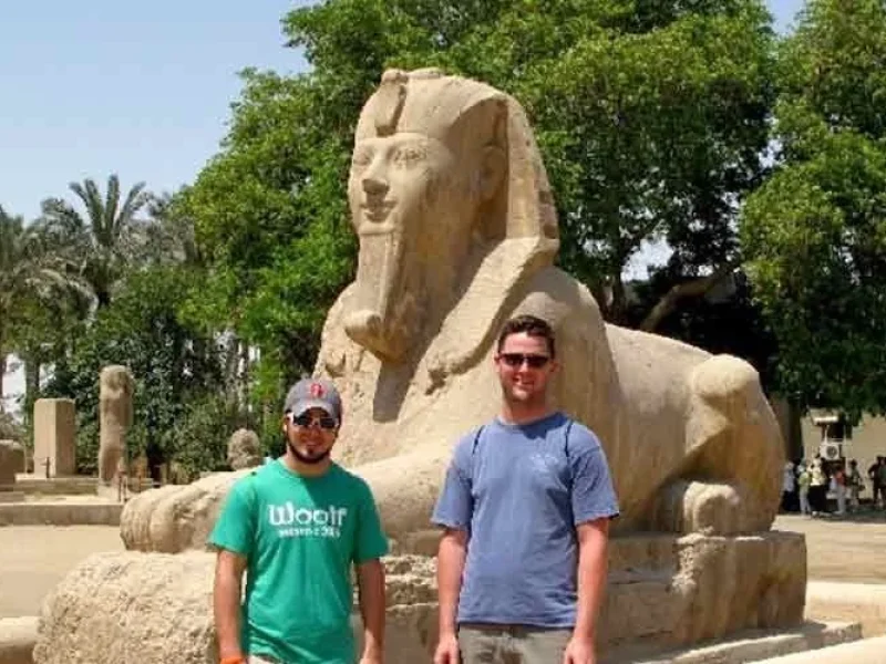 Two tourists standing beside the Great Sphinx at the ancient city of Memphis, Egypt, surrounded by palm trees and archaeological ruins.