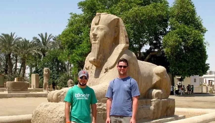 Two tourists standing beside the Great Sphinx at the ancient city of Memphis, Egypt, surrounded by palm trees and archaeological ruins.