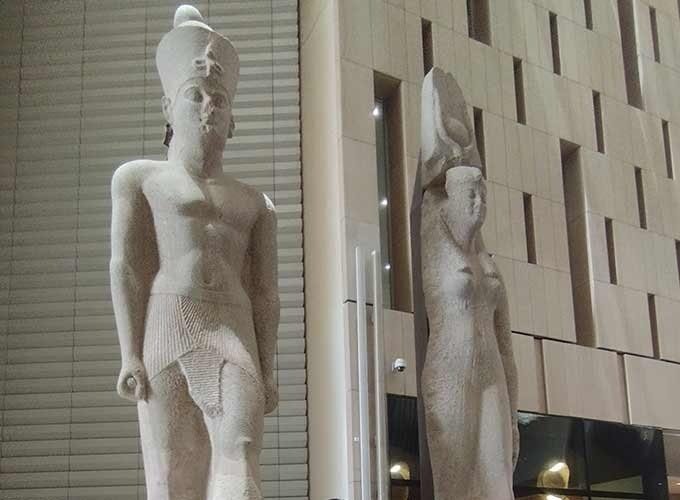 Two towering ancient Egyptian stone statues stand prominently inside a modern hall with light-colored textured walls. The main statue depicts a Pharaonic king wearing a royal kilt and crown, while a smaller female statue stands beside it during a Grand Egyptian Museum trip. The sleek contemporary architecture of the museum interior provides a sharp contrast to the weathered grey stone of these historic artifacts