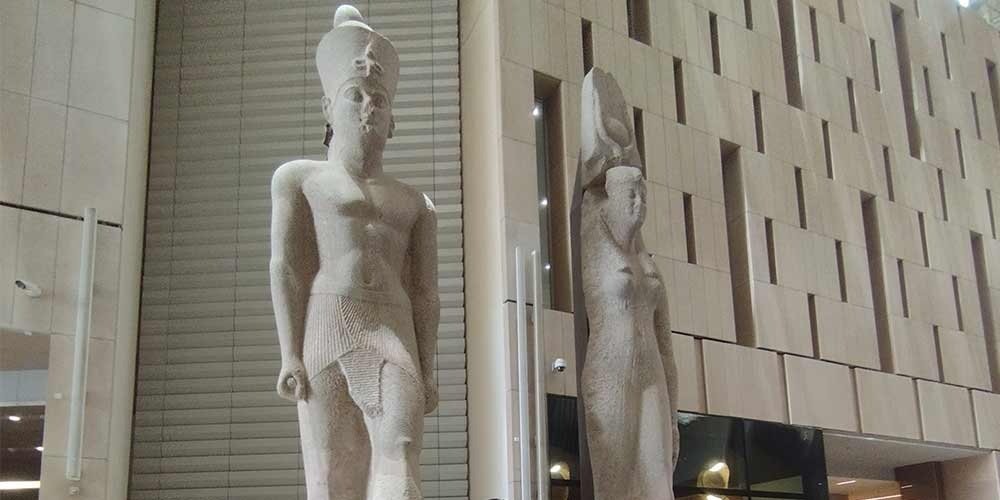 Two towering ancient Egyptian stone statues stand prominently inside a modern hall with light-colored textured walls. The main statue depicts a Pharaonic king wearing a royal kilt and crown, while a smaller female statue stands beside it during a Grand Egyptian Museum trip. The sleek contemporary architecture of the museum interior provides a sharp contrast to the weathered grey stone of these historic artifacts