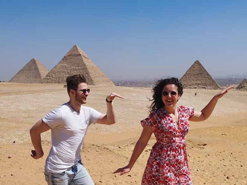 A happy young man in a white t-shirt and a woman in a floral red dress are striking a playful Egyptian pose in the desert. The three Great Pyramids of Giza stand under a clear sky in the background of this Grand Egyptian Museum& tour