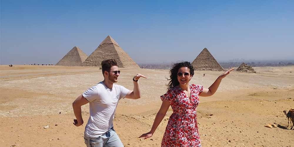 A happy young man in a white t-shirt and a woman in a floral red dress are striking a playful Egyptian pose in the desert. The three Great Pyramids of Giza stand under a clear sky in the background of this Grand Egyptian Museum& tour