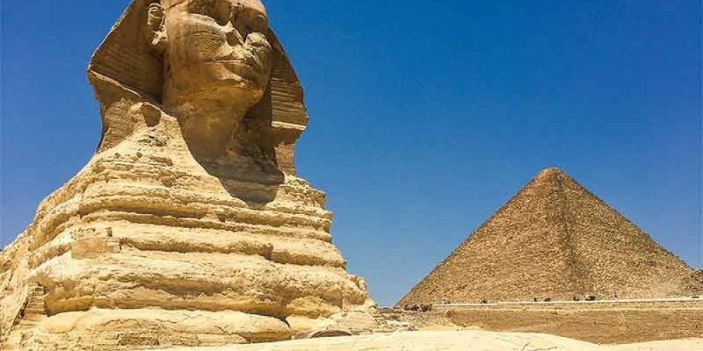 The Great Sphinx of Giza stands in profile with its monumental lion body and human head carved from limestone. A sharp triangular pyramid rises in the background under a deep blue, cloudless sky during a Grand Egyptian Museum& tour