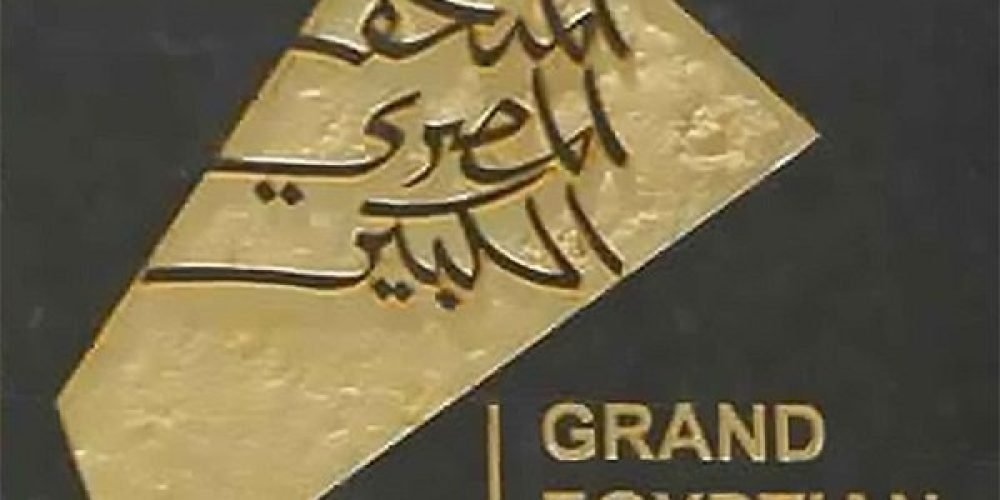 The official logo of the Grand Egyptian Museum& features a gold triangular shape with Arabic calligraphy on a black background. Below the golden emblem, the English name "GRAND EGYPTIAN MUSEUM" is written in clean, gold sans-serif capital letters