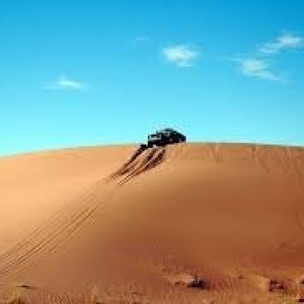 A 4x4 off-road vehicle driving on top of a massive golden sand dune under a clear blue sky in the desert. Adventure scene capturing dune tracks and vast empty desert landscape during Grand Safary in Ghost City