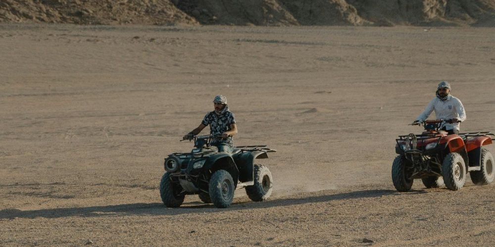 Two men riding quad bikes across a wide sandy desert with rocky mountains in the background under natural daylight. Outdoor adventure scene showing off-road quad biking during Grand Safary in Ghost Cit