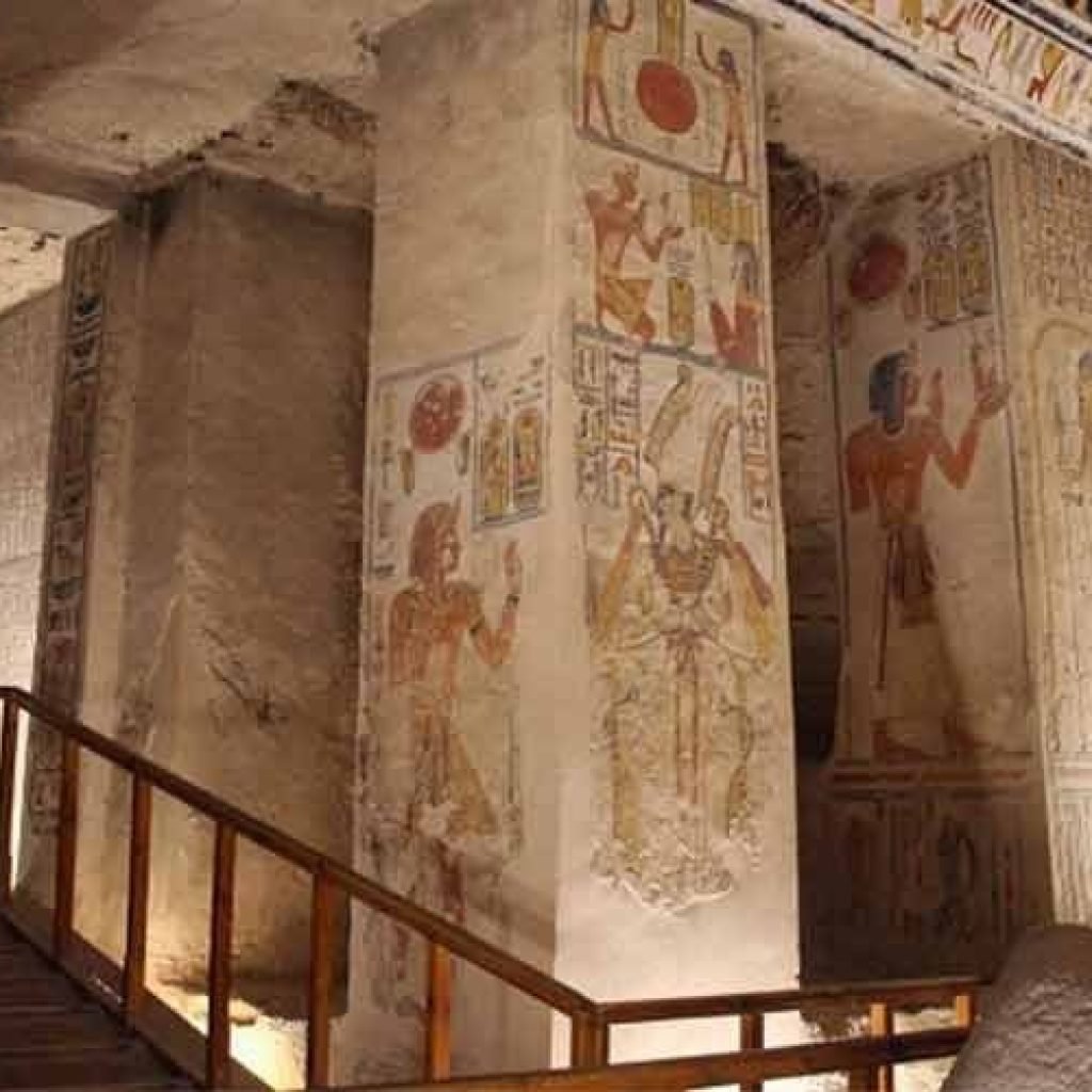 A wide, interior view of an ancient Egyptian royal tomb featuring massive square limestone pillars. Each pillar is decorated with detailed, colorful reliefs and hieroglyphics depicting pharaohs and gods. A wooden walkway with a handrail for visitors leads through the chamber. The ceiling is covered in intricate astronomical paintings and symbols. The walls are filled with horizontal rows of ancient texts and scenes, all illuminated by warm artificial lighting, a key stop on a Group Day Tour to Luxor