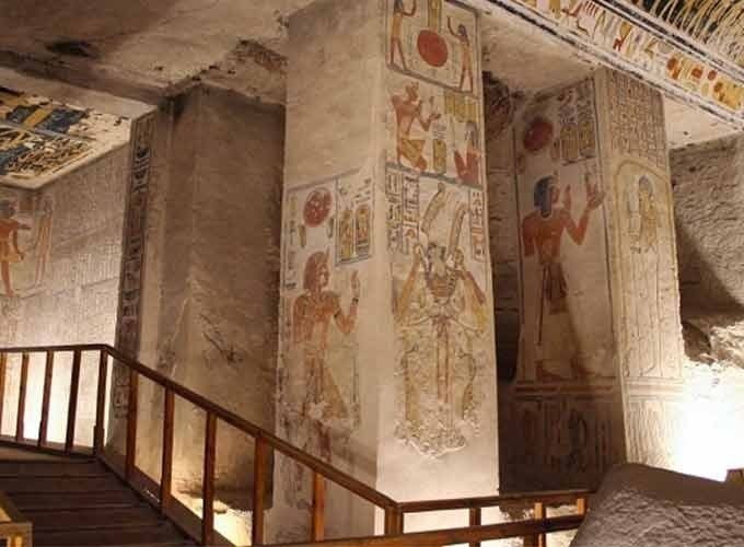 A wide, interior view of an ancient Egyptian royal tomb featuring massive square limestone pillars. Each pillar is decorated with detailed, colorful reliefs and hieroglyphics depicting pharaohs and gods. A wooden walkway with a handrail for visitors leads through the chamber. The ceiling is covered in intricate astronomical paintings and symbols. The walls are filled with horizontal rows of ancient texts and scenes, all illuminated by warm artificial lighting, a key stop on a Group Day Tour to Luxor