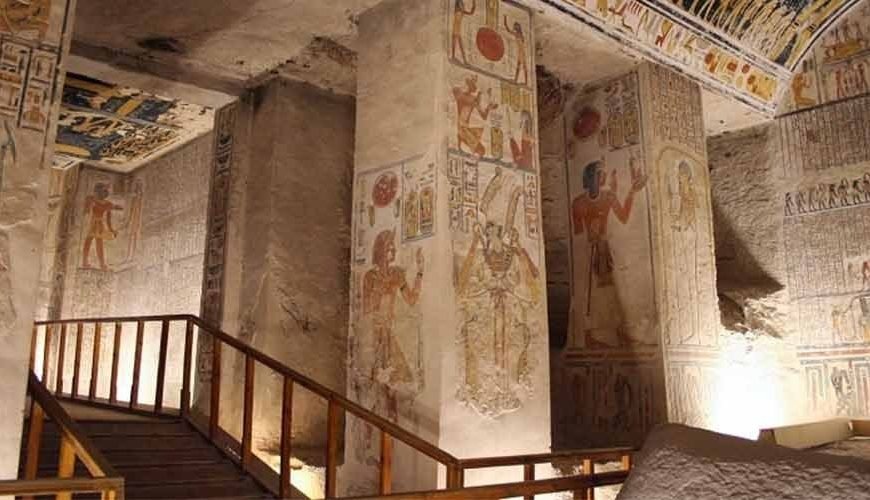 A wide, interior view of an ancient Egyptian royal tomb featuring massive square limestone pillars. Each pillar is decorated with detailed, colorful reliefs and hieroglyphics depicting pharaohs and gods. A wooden walkway with a handrail for visitors leads through the chamber. The ceiling is covered in intricate astronomical paintings and symbols. The walls are filled with horizontal rows of ancient texts and scenes, all illuminated by warm artificial lighting, a key stop on a Group Day Tour to Luxor