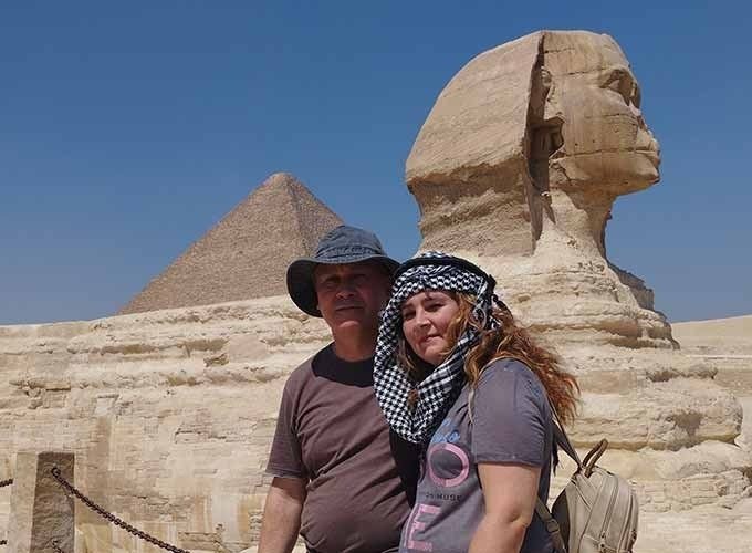 A couple posing for a photo in front of the Great Sphinx of Giza and a pyramid during their Low Budget Egypt Tour. The man wears a sun hat and the woman has a checkered scarf over her head, with the ancient limestone monuments standing tall under a clear blue sky
