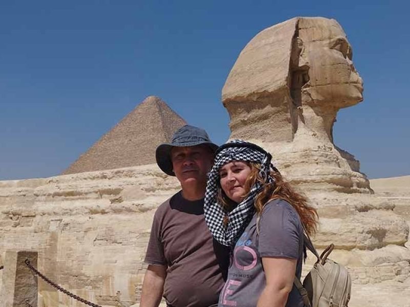 A couple posing for a photo in front of the Great Sphinx of Giza and a pyramid during their Low Budget Egypt Tour. The man wears a sun hat and the woman has a checkered scarf over her head, with the ancient limestone monuments standing tall under a clear blue sky