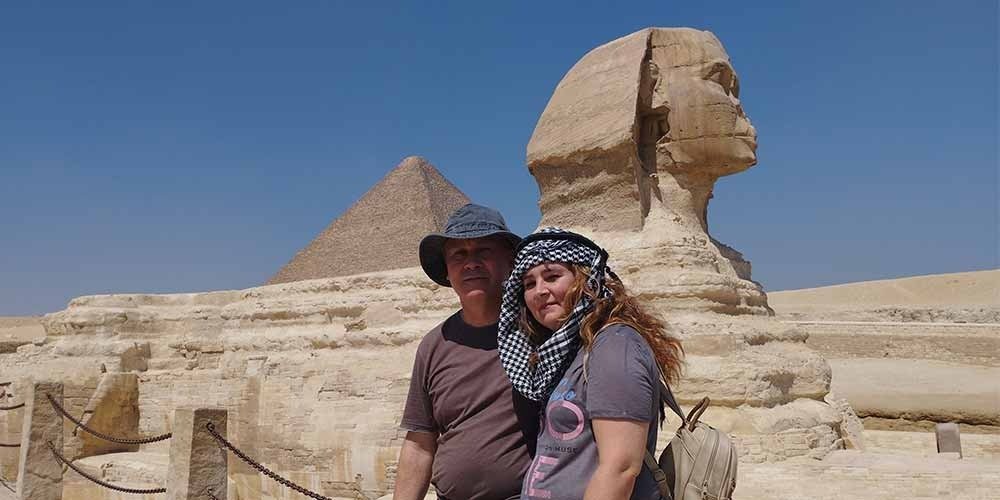 A couple posing for a photo in front of the Great Sphinx of Giza and a pyramid during their Low Budget Egypt Tour. The man wears a sun hat and the woman has a checkered scarf over her head, with the ancient limestone monuments standing tall under a clear blue sky