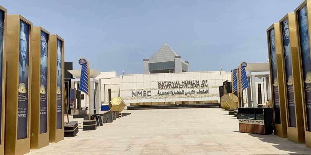 The main entrance of the National Museum of Egyptian Civilization (NMEC) featuring tall golden banners and modern white architecture during a Low Budget Egypt Tour. A paved courtyard leads toward the museum building with a pyramid-shaped roof and large gold geometric decorations under a clear sky