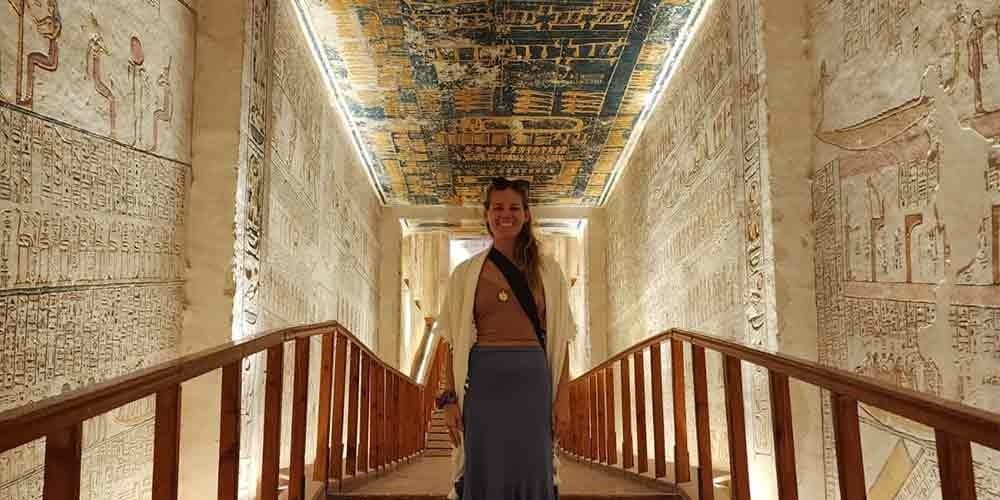 A female tourist standing on a wooden walkway inside a brightly lit ancient Egyptian tomb during a Low Budget Egypt Tour. The walls and high ceiling are completely covered in detailed colorful hieroglyphics and astronomical scenes under warm artificial lighting.