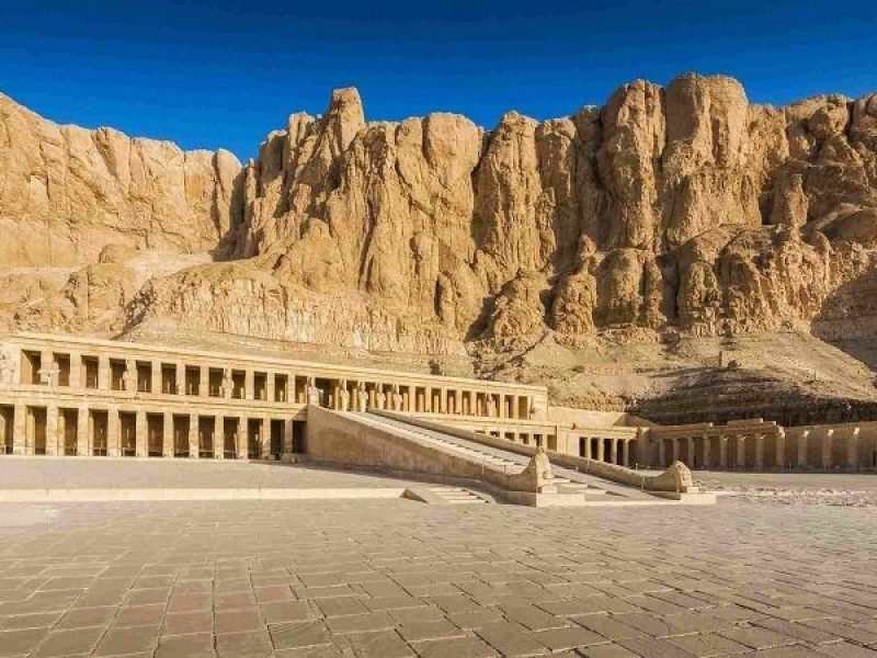A panoramic view of the Mortuary Temple of Hatshepsut featuring its grand colonnaded terraces and central ramp built against massive limestone cliffs. The vast paved courtyard in the foreground leads toward the ancient monumental structure under a clear, deep blue sky during a Luxor Tour from Safaga Port
