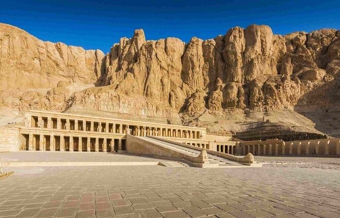 A panoramic view of the Mortuary Temple of Hatshepsut featuring its grand colonnaded terraces and central ramp built against massive limestone cliffs. The vast paved courtyard in the foreground leads toward the ancient monumental structure under a clear, deep blue sky during a Luxor Tour from Safaga Port