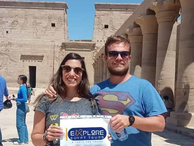 A smiling couple poses in the courtyard of the Temple of Philae in Aswan. The woman is on the left wearing a grey t-shirt and sunglasses, and the man is on the right in a blue t-shirt with a Superman logo. They are holding a white sign that says 