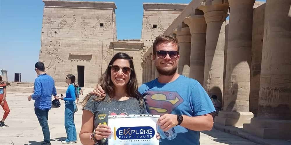A smiling couple poses in the courtyard of the Temple of Philae in Aswan. The woman is on the left wearing a grey t-shirt and sunglasses, and the man is on the right in a blue t-shirt with a Superman logo. They are holding a white sign that says "EXPLORE EGYPT TOURS". In the background, ancient sandstone pylons with carved Egyptian deities are visible under a clear blue sky, alongside a row of large stone columns. This visit often follows a VIP tour inside the Great Pyramid.