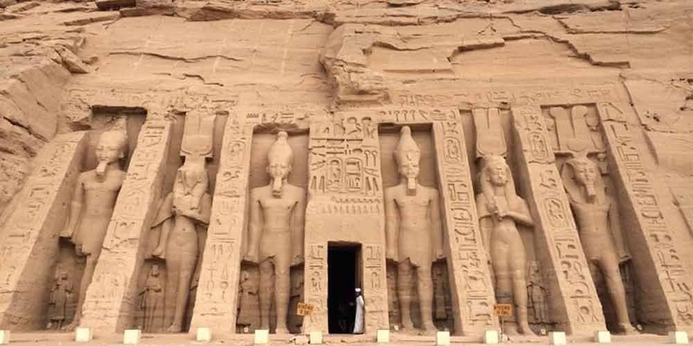 A detailed wide shot of the rock-cut facade of the Small Temple of Nefertari at Abu Simbel. The facade features six large standing statues carved into the mountain: four of Pharaoh Ramesses II and two of Queen Nefertari, each standing between deep buttresses decorated with vertical rows of hieroglyphics. A dark rectangular entrance is centered at the bottom, with a person in white clothing standing in front. The surrounding rock is rough and light brown, typical of a Nile Cruise trip stop in Nubia.