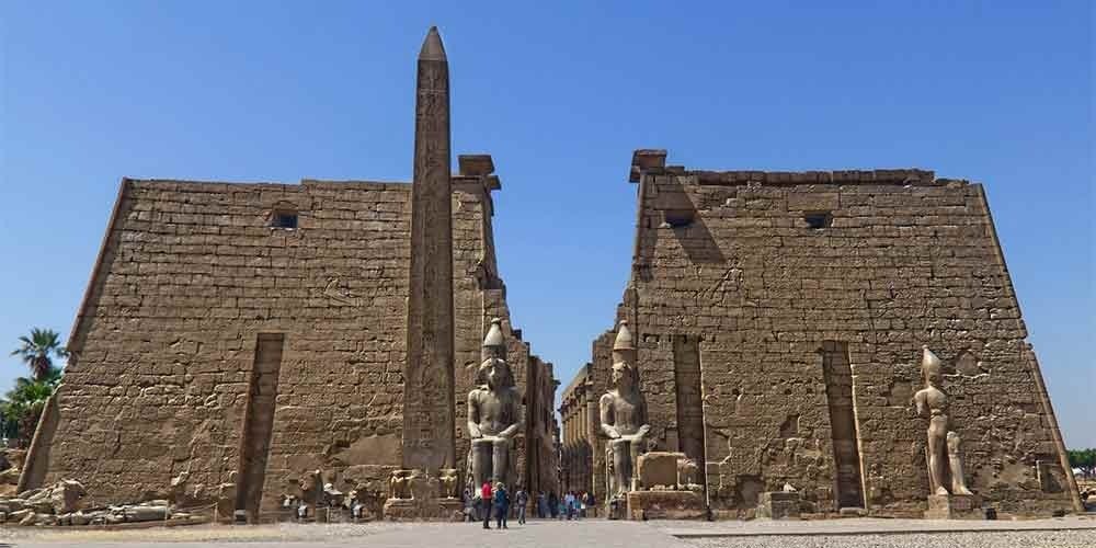 A majestic wide shot of the entrance to Luxor Temple in Egypt. The facade features two massive ancient stone pylons with detailed carvings of pharaohs. In front of the left pylon stands a tall, slender pink granite obelisk covered in hieroglyphs. Two colossal seated statues of Ramesses II guard the central gateway, while a smaller standing statue is visible on the far right. A clear blue sky and a few tourists at the base highlight the scale of this stop during a Nile Cruise trip.