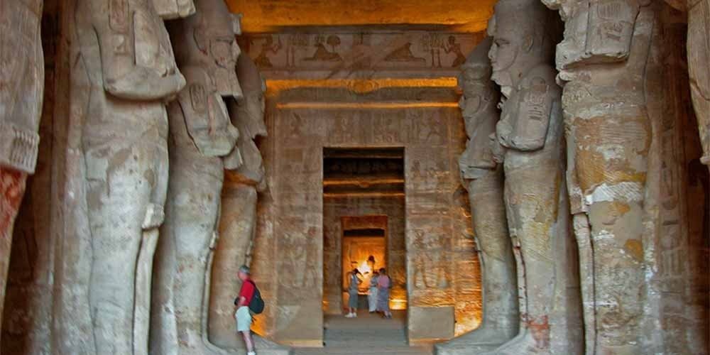 An interior shot of the Great Temple of Abu Simbel featuring the Hypostyle Hall. Two rows of colossal statues of Ramesses II in the form of Osiris stand against pillars, flanking a central aisle that leads to the inner sanctuary. The statues are carved with crossed arms and tall crowns. The walls and ceiling are covered in detailed ancient Egyptian reliefs and hieroglyphics, bathed in warm yellow artificial lighting. A few tourists are visible in the distance near the sanctuary during their Nile Cruise trip