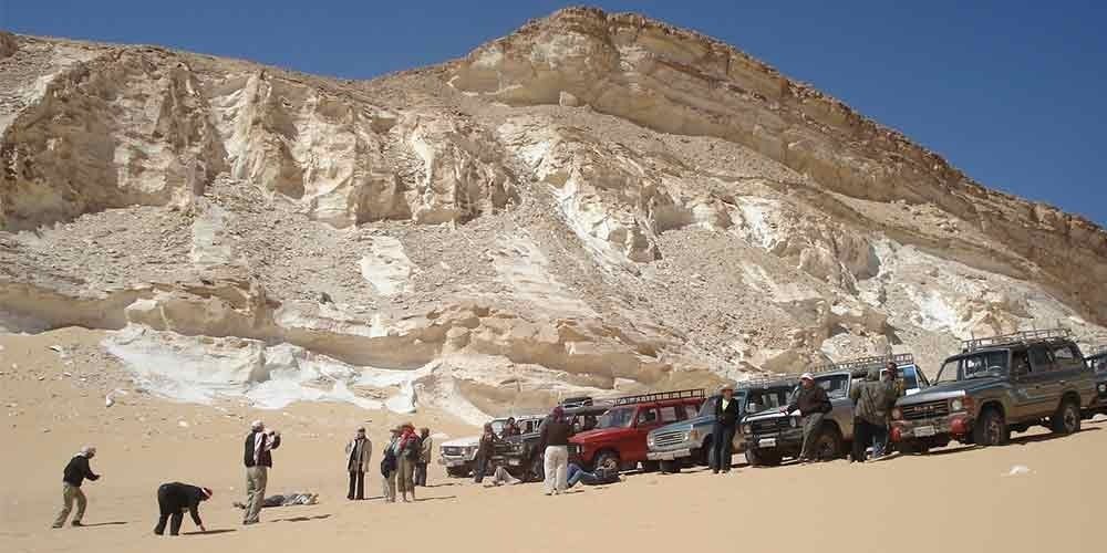 Grupo de seis turistas posando no Deserto Branco, com céu parcialmente nublado e formações rochosas ao fundo; uma mulher segura uma placa da Explore Egypt Tours.