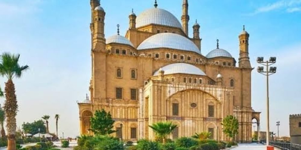 The majestic Mosque of Muhammad Ali in Cairo with its high minarets and large silver domes, surrounded by green gardens and palm trees under a bright blue sky