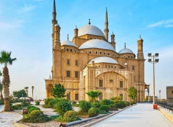 The majestic Mosque of Muhammad Ali in Cairo with its high minarets and large silver domes, surrounded by green gardens and palm trees under a bright blue sky