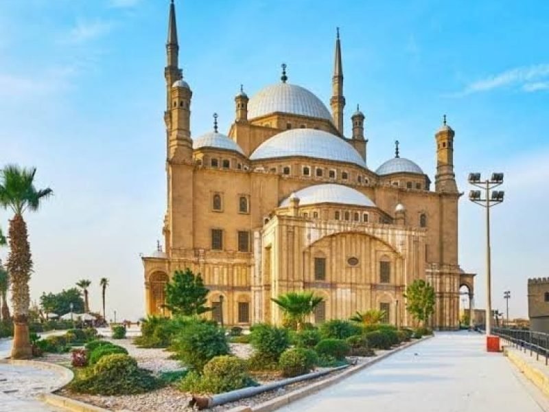 The majestic Mosque of Muhammad Ali in Cairo with its high minarets and large silver domes, surrounded by green gardens and palm trees under a bright blue sky