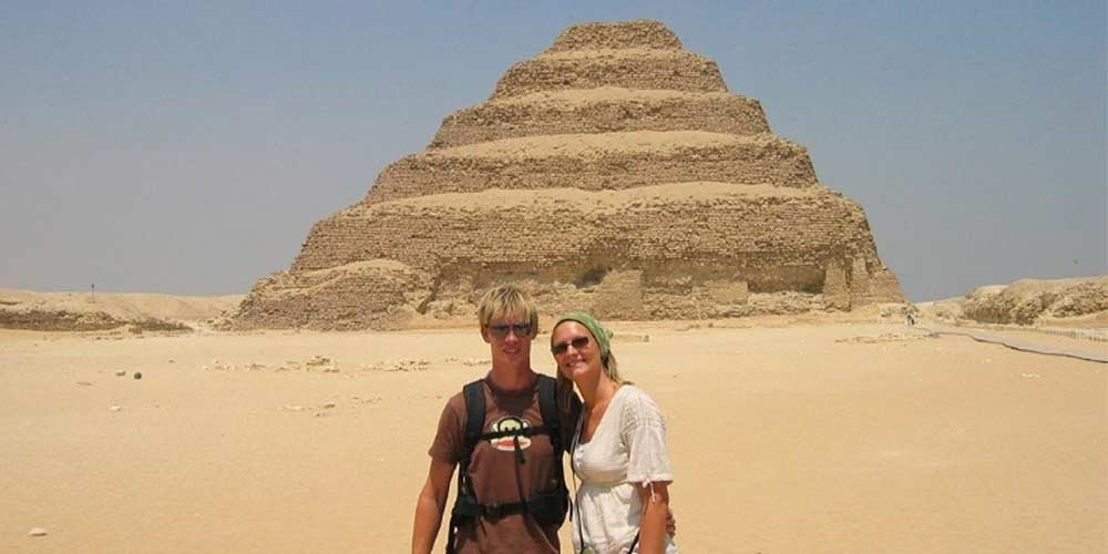 A man in a brown t-shirt and a woman in a white top and green headscarf stand together in the desert sands with the ancient Step Pyramid of Djoser behind them