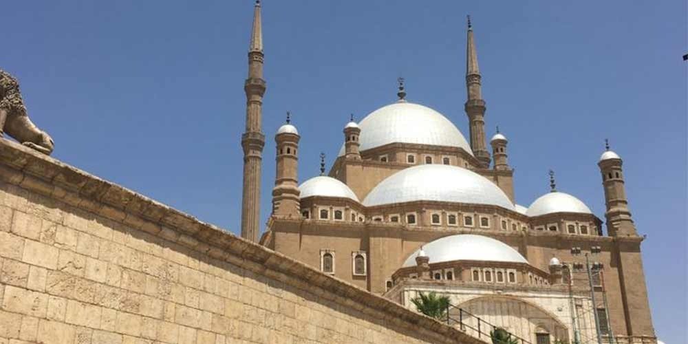 A detailed low-angle shot of the Mosque of Muhammad Ali during an Overnight excursion, showing the large silver domes, slender minarets, and the sturdy stone perimeter wall against a clear blue sky